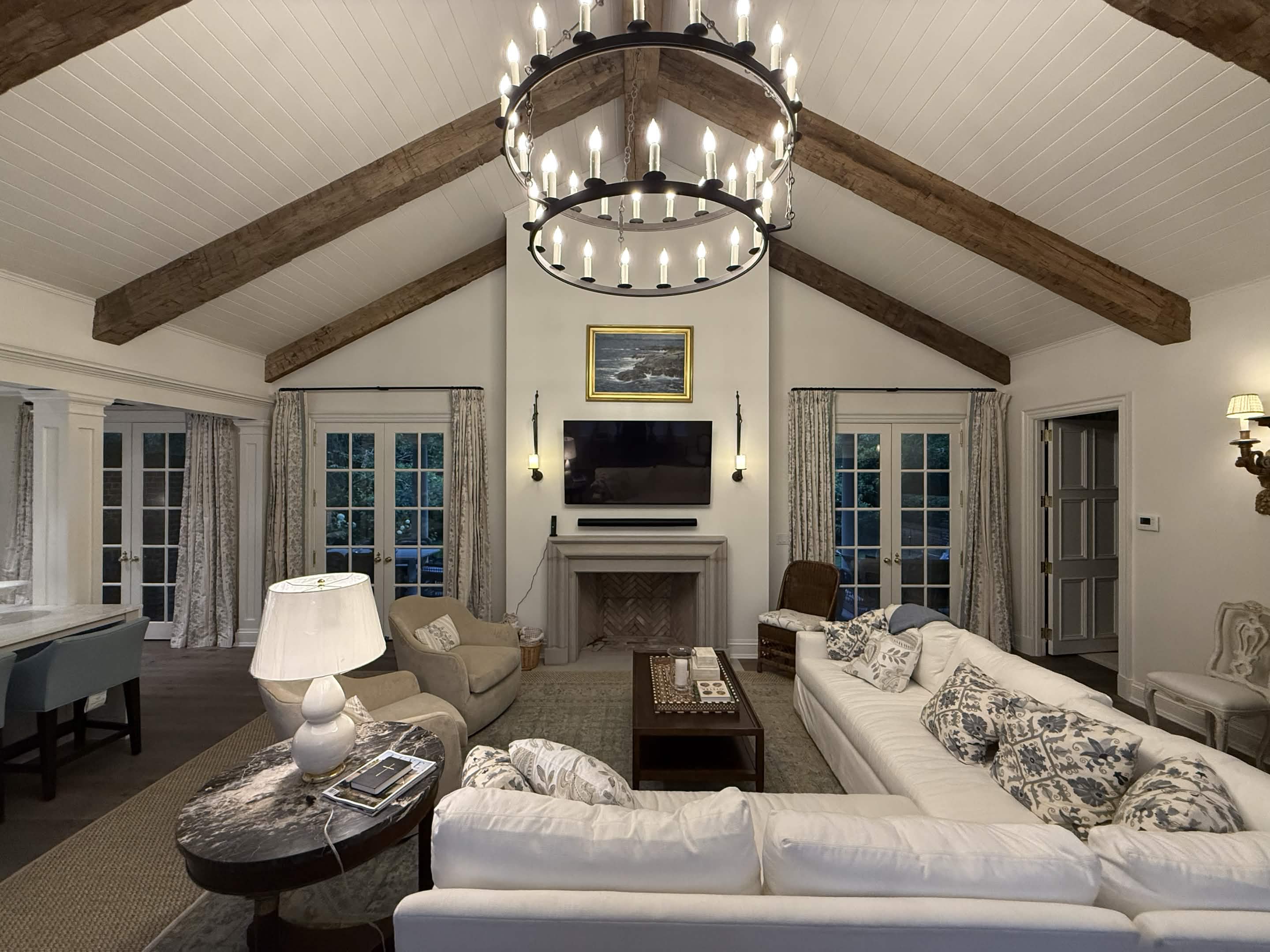 A vaulted great room with hand-hewn timber beams, an iron chandelier, and a stone fireplace at dusk